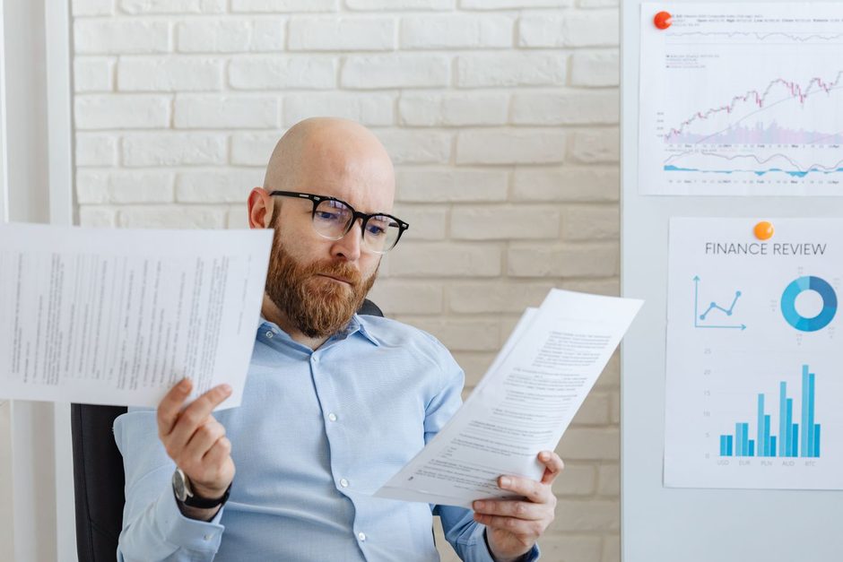 Photo by https://kaboompics.com/ on Pexels Man in a blue shirt reviewing printed analytics papers beside a whiteboard chart. Photo by https://kaboompics.com/ on Pexels