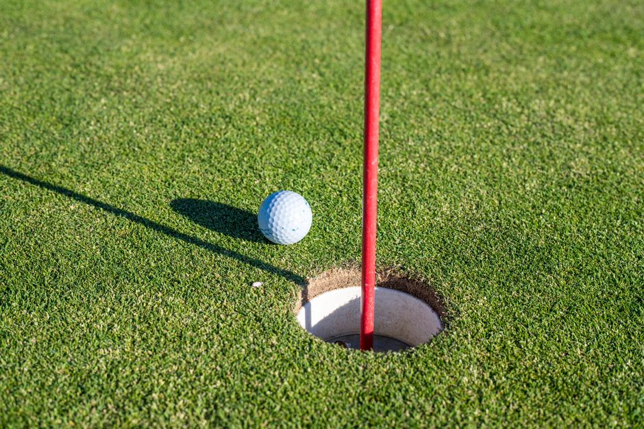 A close-up of a golf ball near a hole on a green with a red flagpole, symbolizing precision and sport. Photo by Mateusz Feliksik on Pexels