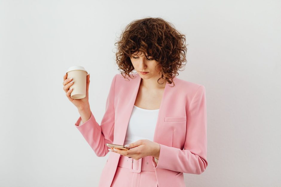 Woman in a pink blazer holding a smartphone against a plain bright wall. Photo by https://kaboompics.com/ on Pexels