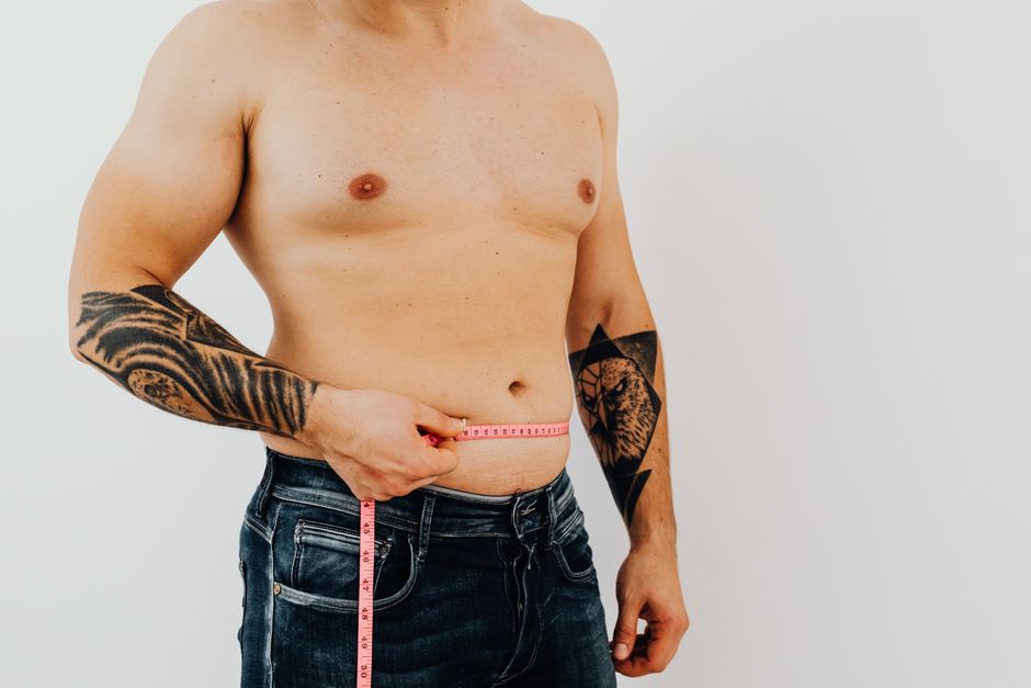 Shirtless man using measuring tape to check waist size, showcasing tattoos for fitness concept. Photo by https://kaboompics.com/ on Pexels
