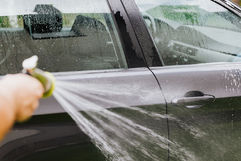 A person uses a hose to wash a black car outdoors, creating water splashes. Photo by https://kaboompics.com/ on Pexels