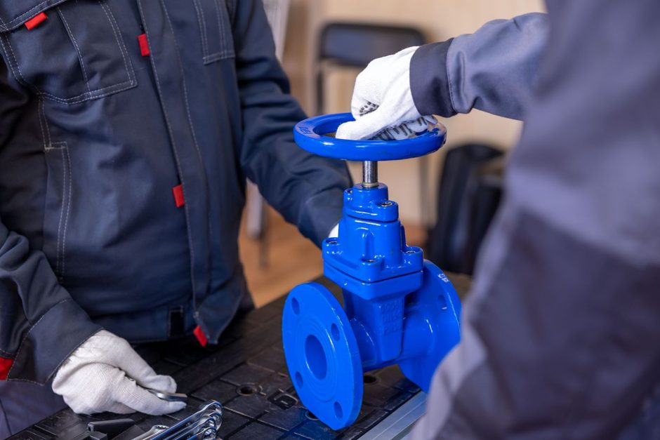 Plumber in blue overalls turning a large blue valve on a pipe. Photo by Sergei Starostin on Pexels