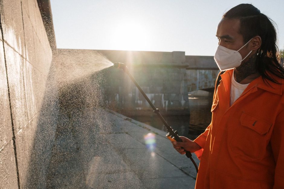Worker in an orange suit and mask pressure washing a patio in sunlight. Photo by cottonbro studio on Pexels