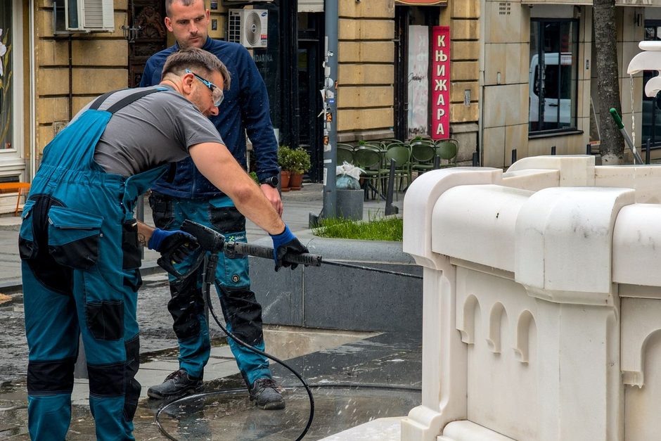 Two workers pressure washing a pavement outside a building. Photo by Max Grakov on Pexels