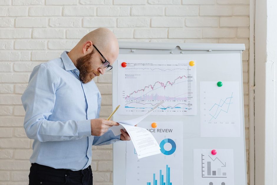 Photo by https://kaboompics.com/ on Pexels Analyst standing at a whiteboard with charts while reviewing a printed report. Photo by https://kaboompics.com/ on Pexels
