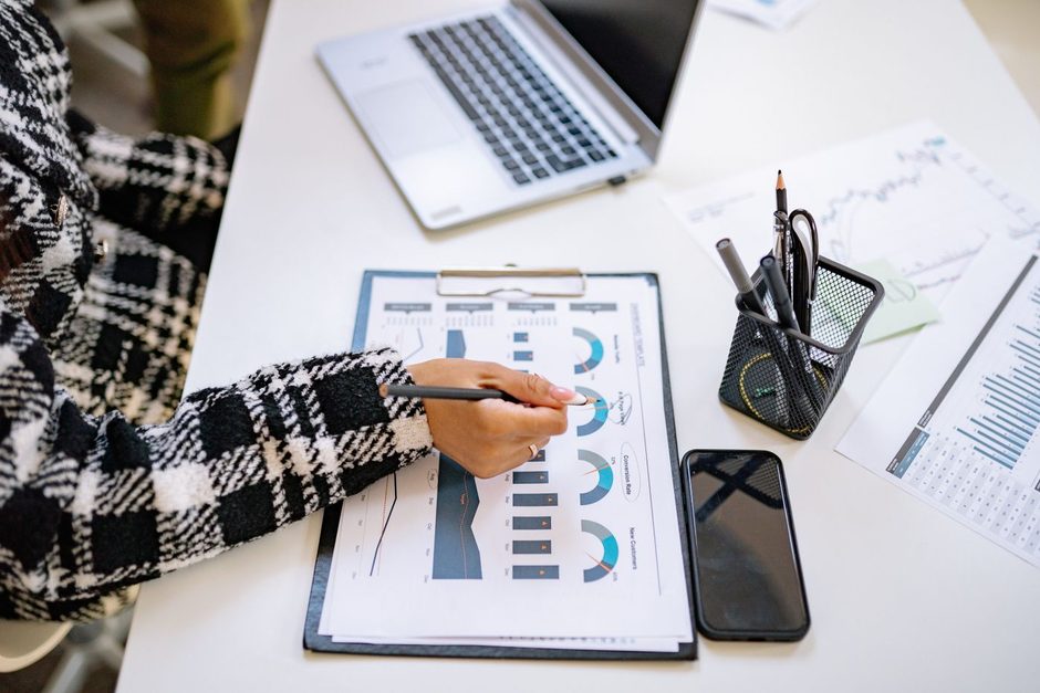 Hand pointing at printed charts beside a laptop on a white desk. Photo by Yan Krukau on Pexels