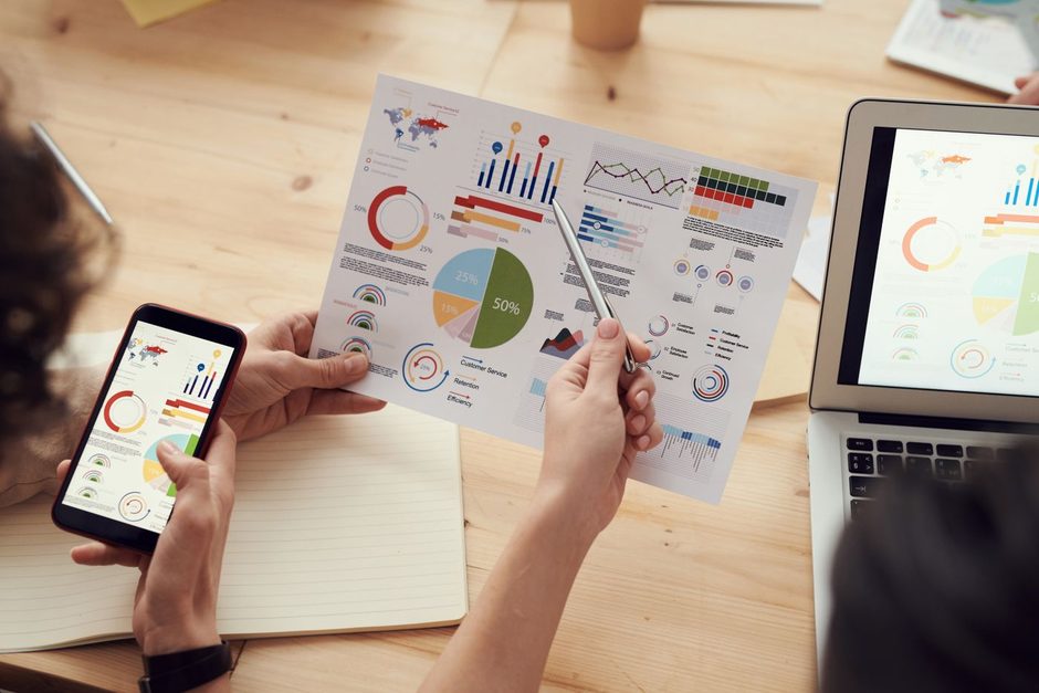 Hands holding a phone over printed charts while reviewing social ad results. Photo by fauxels on Pexels