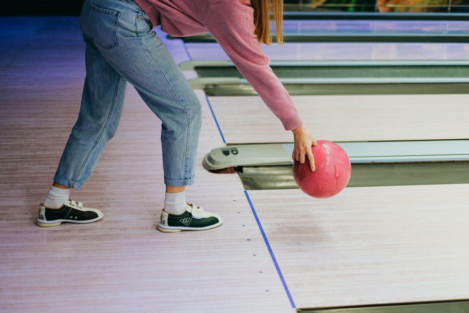 A woman in jeans and sweater playing bowling, throwing a pink ball down the lane indoors. Photo by Anna Shvets on Pexels