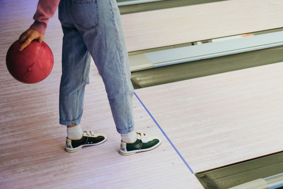 A person holding a pink bowling ball in a colorful bowling alley wearing green bowling shoes. Photo by Anna Shvets on Pexels