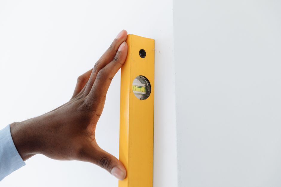 A close-up of a hand using a yellow level tool against a white wall to ensure vertical alignment. Photo by Thirdman on Pexels