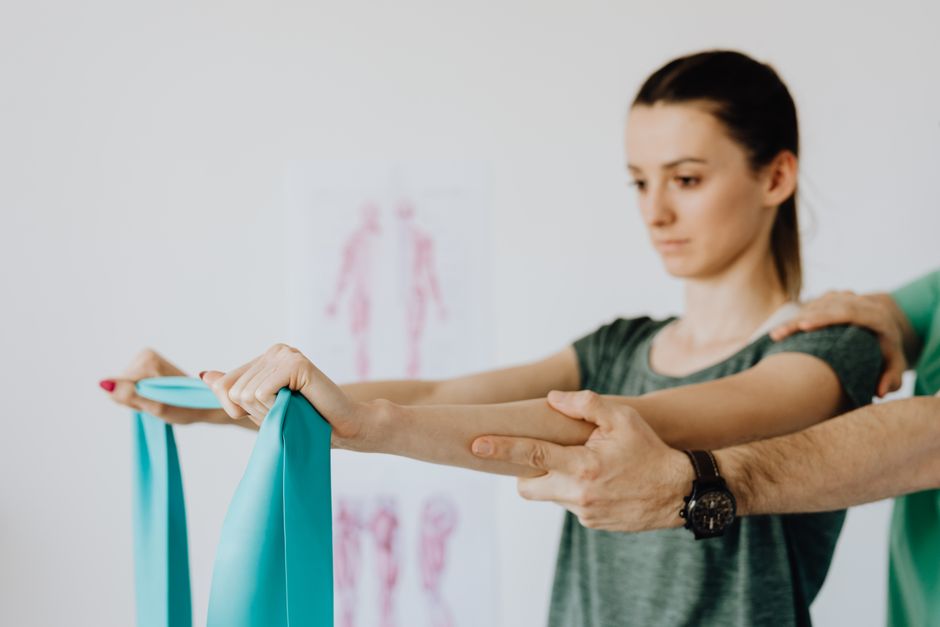 Photo by https://kaboompics.com/ on Pexels Slim young female in casual wear standing with reached arms and elastic tape during medical checkup by crop unrecognizable osteopath in wristwatch. Photo by https://kaboompics.com/ on Pexels