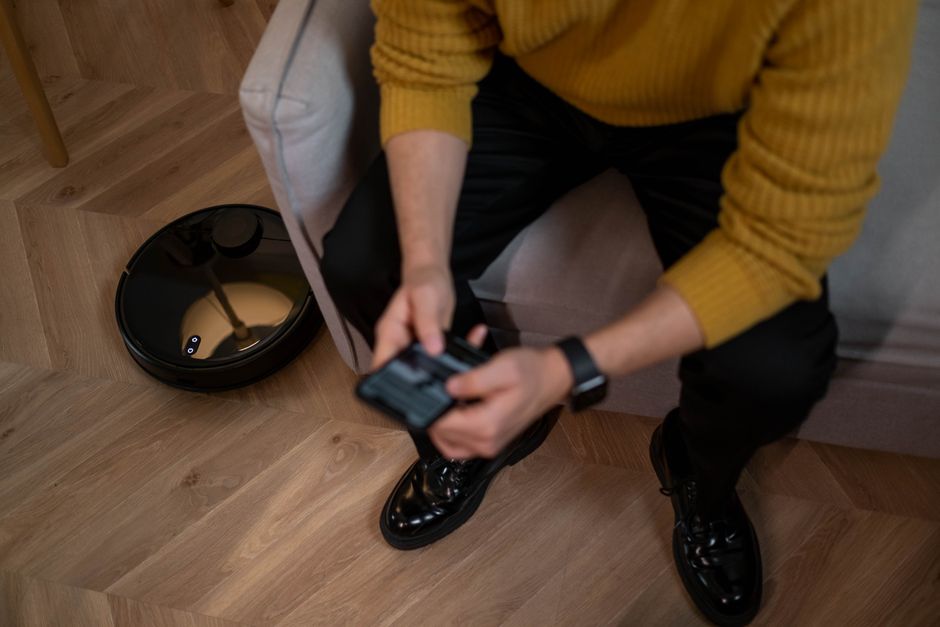 Man in yellow sweater using remote to control robotic vacuum on wooden floor. Photo by cottonbro studio on Pexels