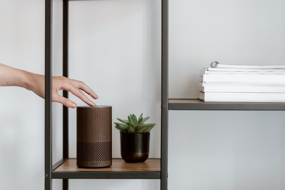 A hand interacts with a smart speaker on a minimalist shelf, featuring a plant and stacked books. Photo by Anete Lusina on Pexels