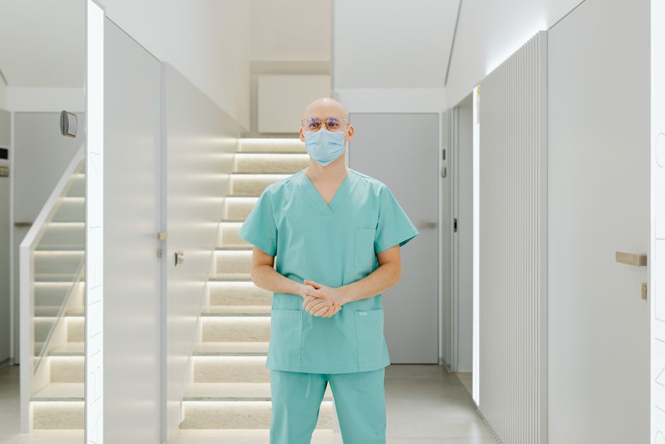 Photo by https://kaboompics.com/ on Pexels Healthcare worker in scrub suit standing indoors with face mask in clinic hallway. Photo by https://kaboompics.com/ on Pexels