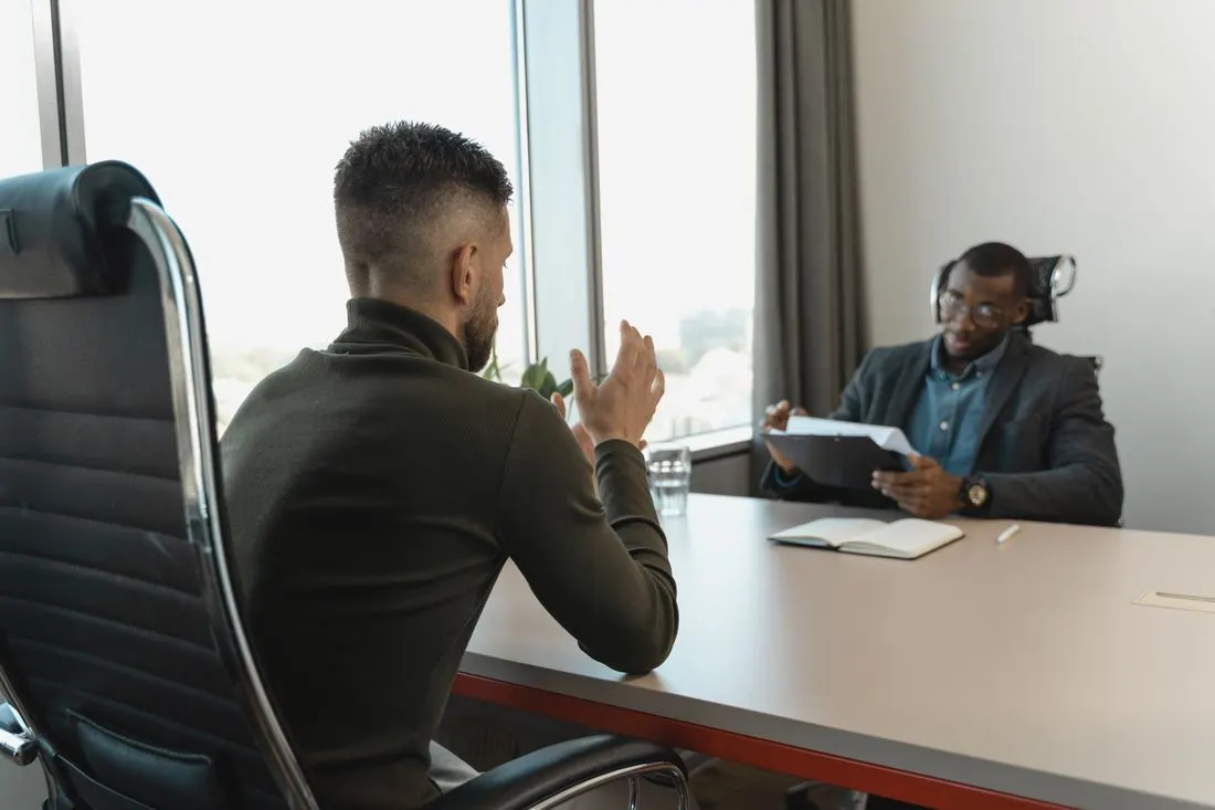 Two men in a meeting across a conference table by a window, one holding a tablet. Photo by Tima Miroshnichenko on Pexels