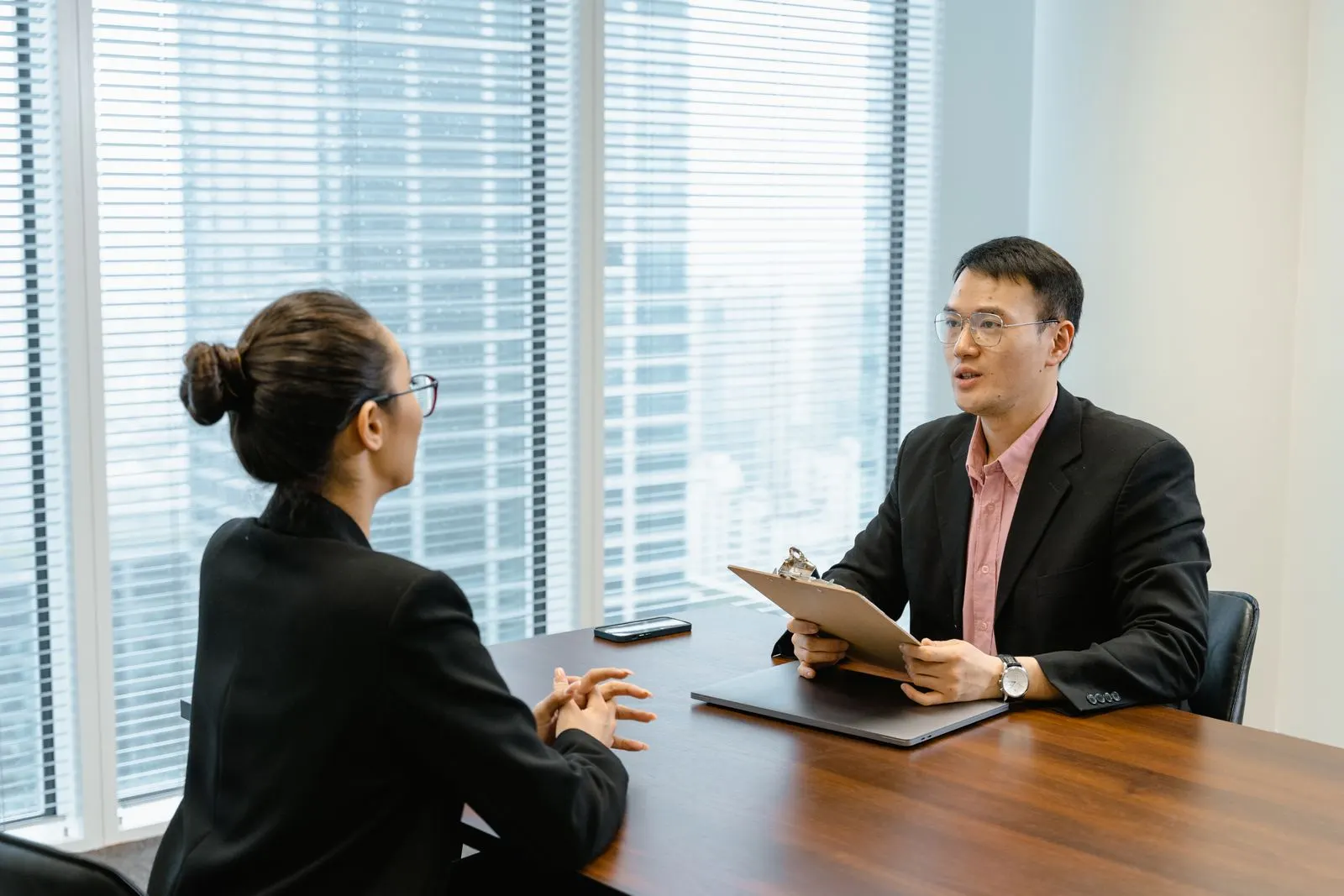 Man in glasses holding a clipboard while talking with a woman across an office desk. Photo by MART PRODUCTION on Pexels