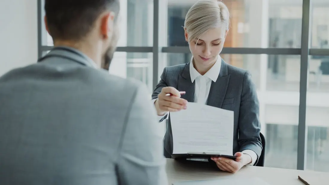 Woman in a grey suit reading a document on a clipboard across a desk from a man. Photo by Vitaly Gariev on Pexels