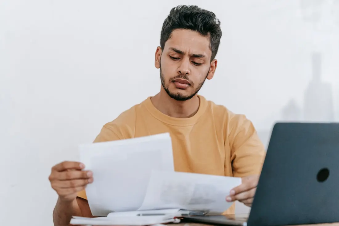 A man at a desk reading printed pages next to a laptop. Photo by Michael Burrows on Pexels