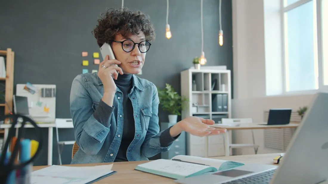 Woman in glasses taking a phone call at a desk with a notebook and laptop. Photo by Vitaly Gariev on Pexels