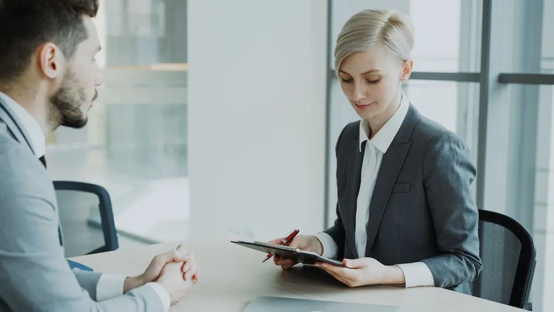 Woman in a grey suit taking notes on a clipboard across a desk from a man. Photo by Vitaly Gariev on Pexels