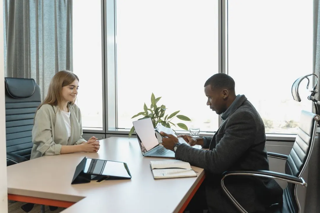 Two people meeting at a desk with a laptop and notebooks by a window. Photo by Tima Miroshnichenko on Pexels