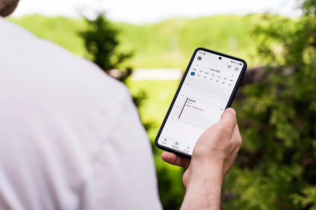 A person using a sauna booking app on a smartphone outdoors, surrounded by greenery. Photo by HUUM  │sauna heaters on Pexels