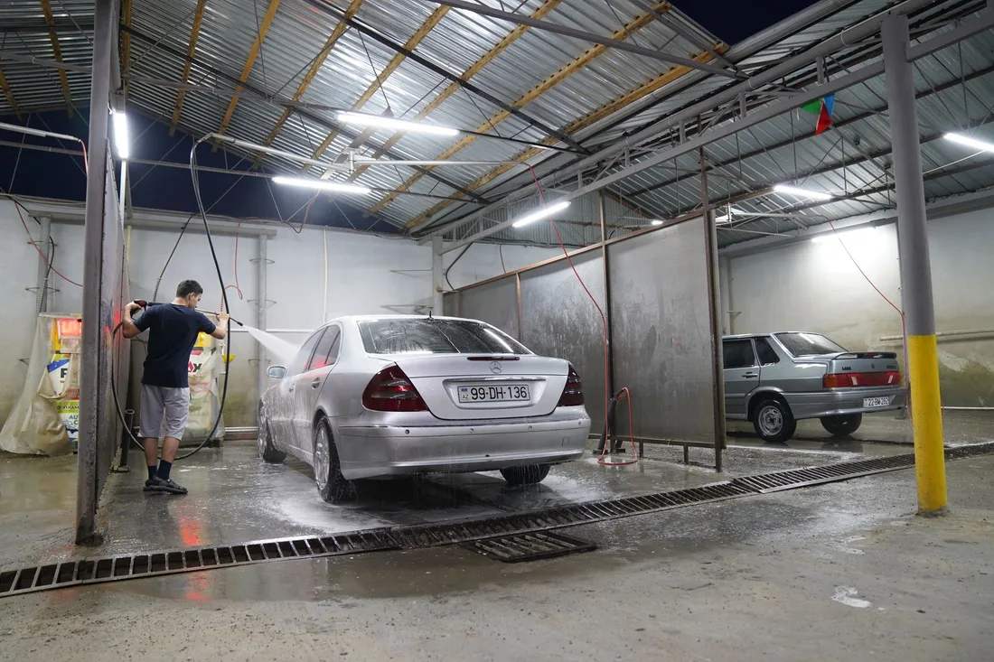 Man washing a silver car with a pressure hose inside a bright car wash bay. Photo by Tahir Xəlfəquliyev on Pexels