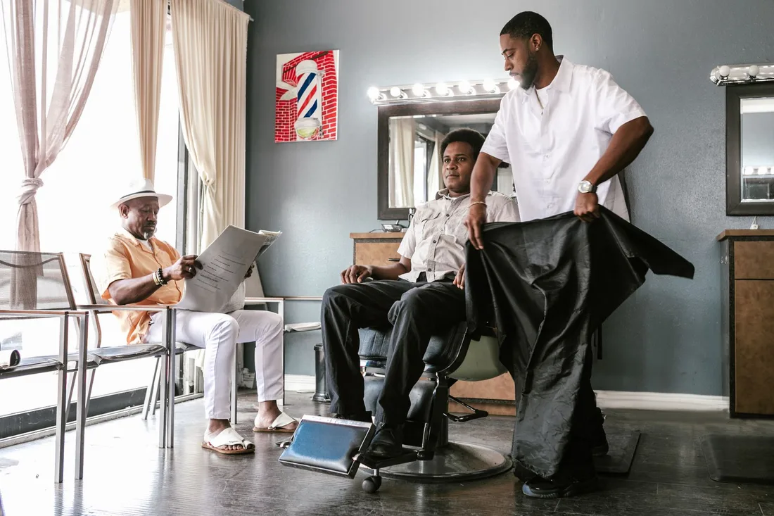 A man receives a haircut in a barber shop while another customer reads a newspaper. Photo by RDNE Stock project on Pexels