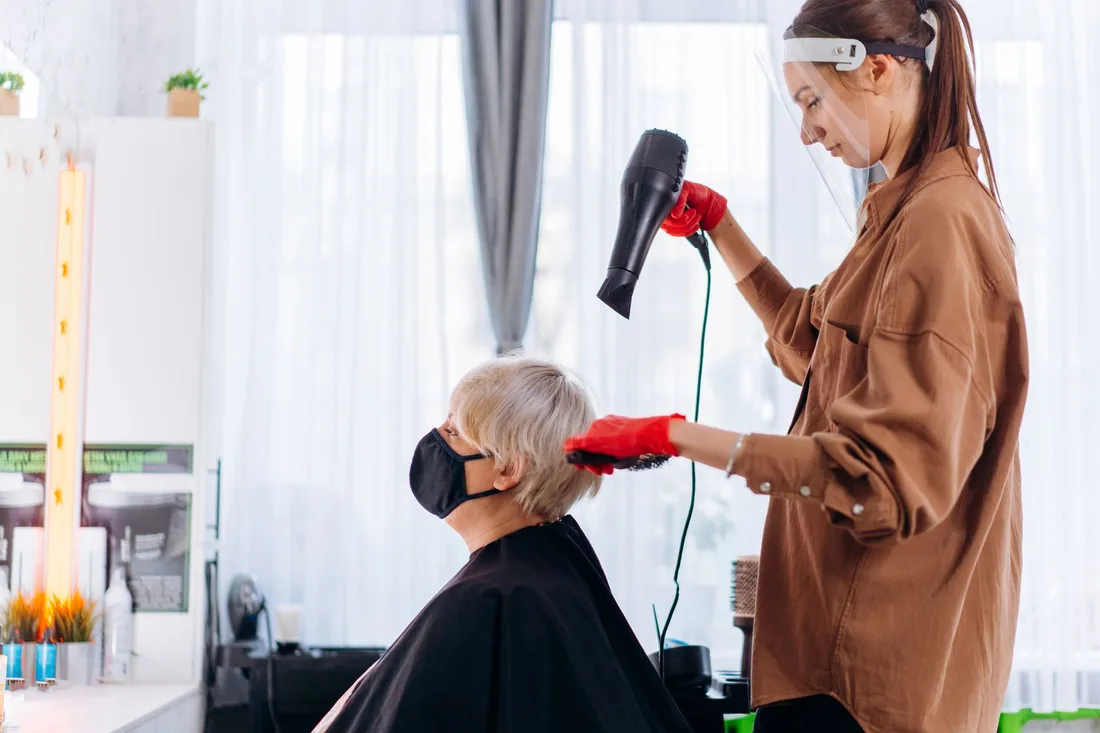 Side view of female hairdresser in protective mask and gloves drying hair of elderly customer while working in salon. Photo by Mikhail Nilov on Pexels