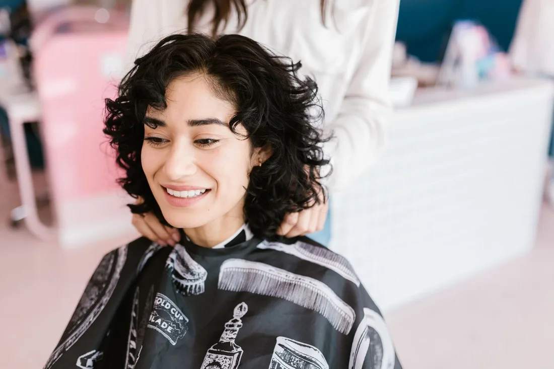 Smiling woman with curly dark hair wearing a salon cape in a bright hair salon. Photo by RDNE Stock project on Pexels
