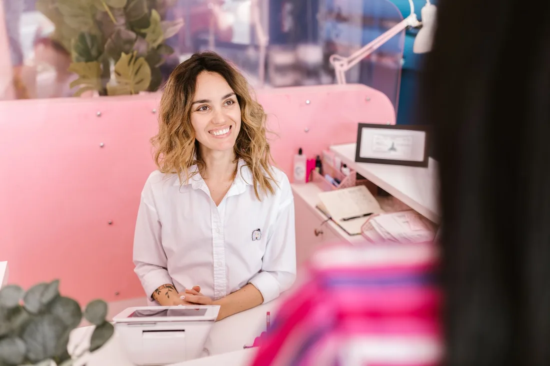 Smiling receptionist at a salon front desk, welcoming clients with a friendly demeanor. Photo by RDNE Stock project on Pexels
