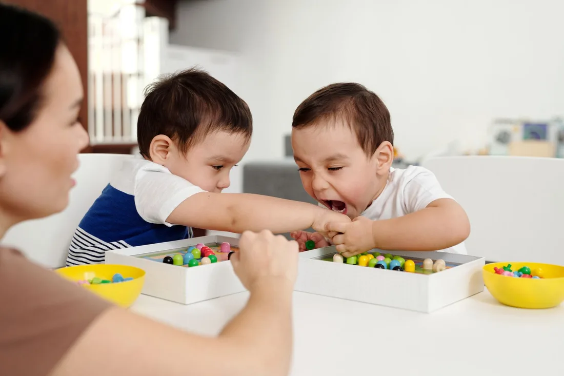 Woman helping two toddlers play with colourful sorting toys at a table. Photo by Jep Gambardella on Pexels