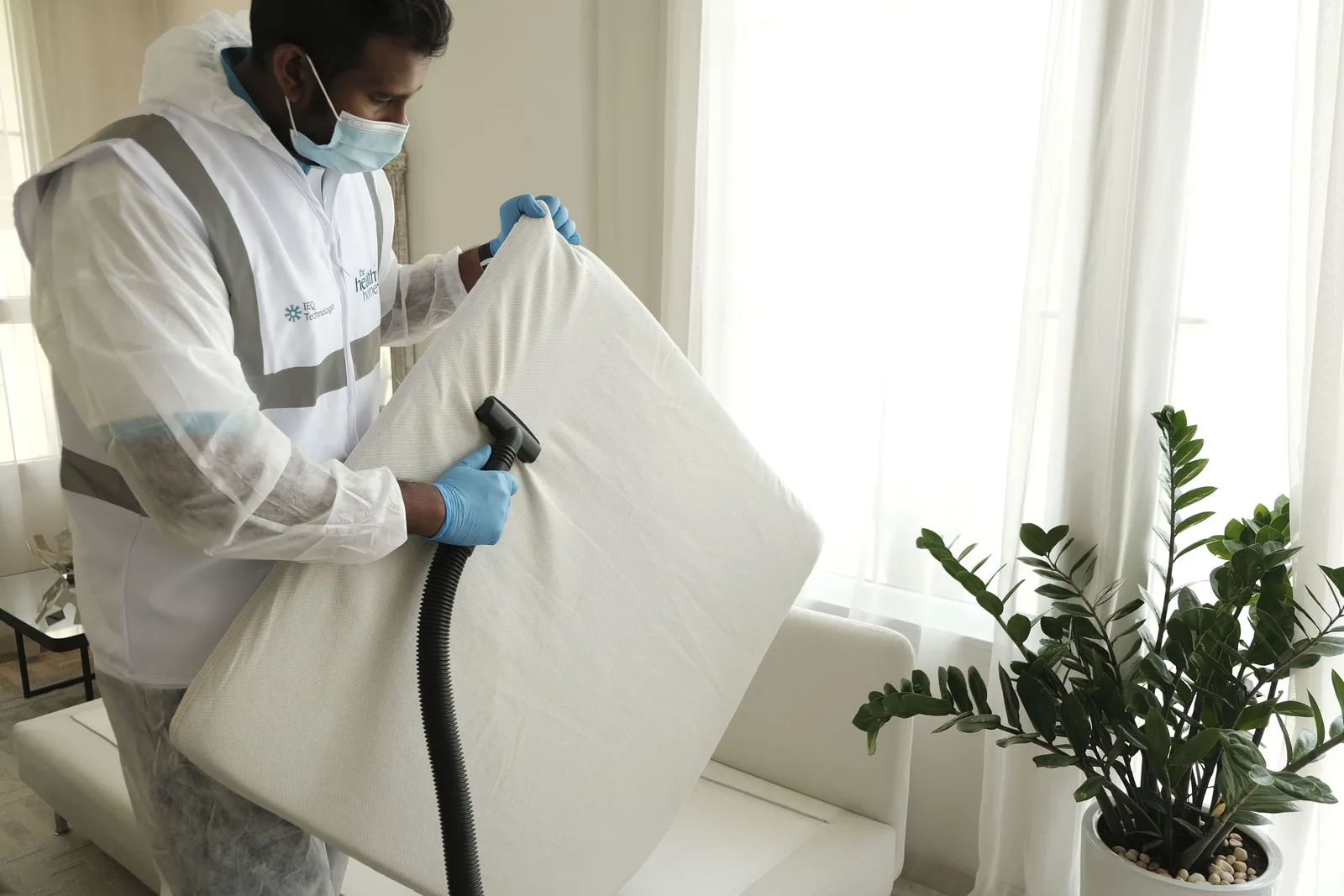 Technician in protective gear and mask holding a white mattress in a bright room. Photo by شركة تعقيم بالرياض thehealthyhome.me/ar/sa on Pexels
