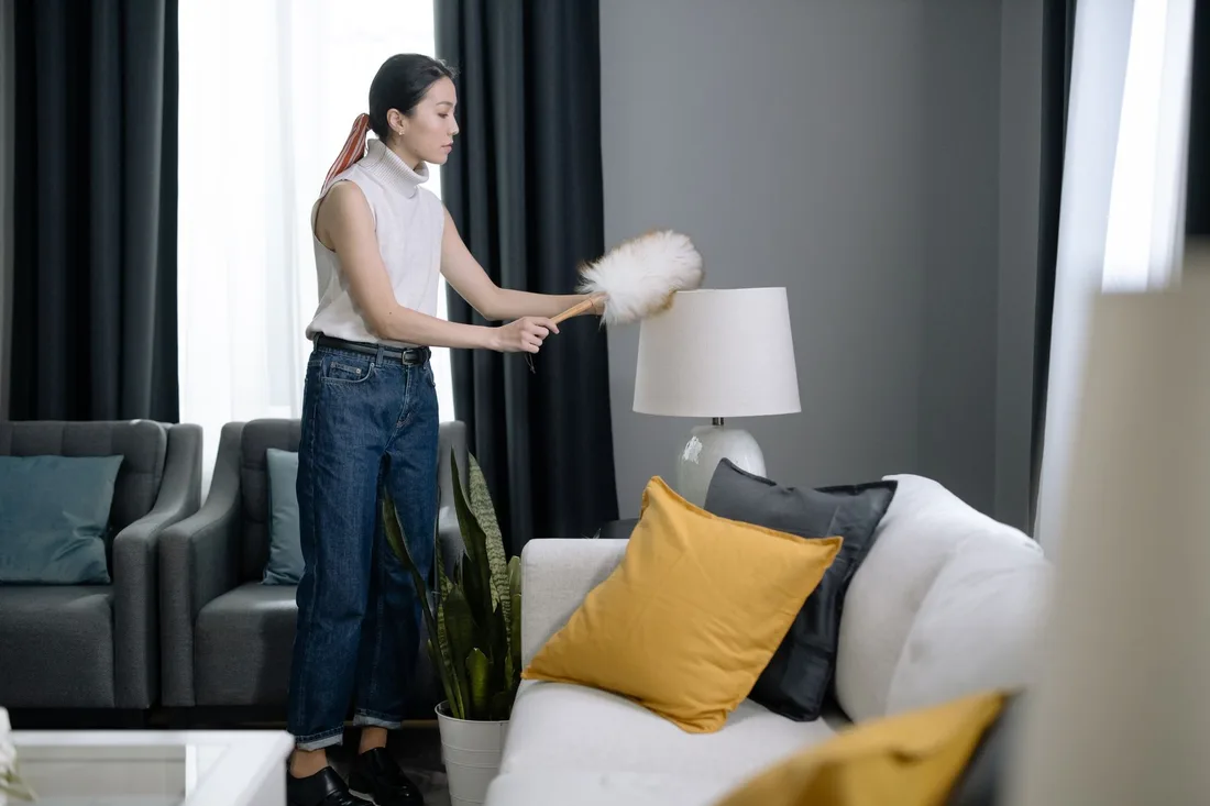 Woman dusting a tidy living room with sofa and cushions in Australia. Photo by cottonbro studio on Pexels