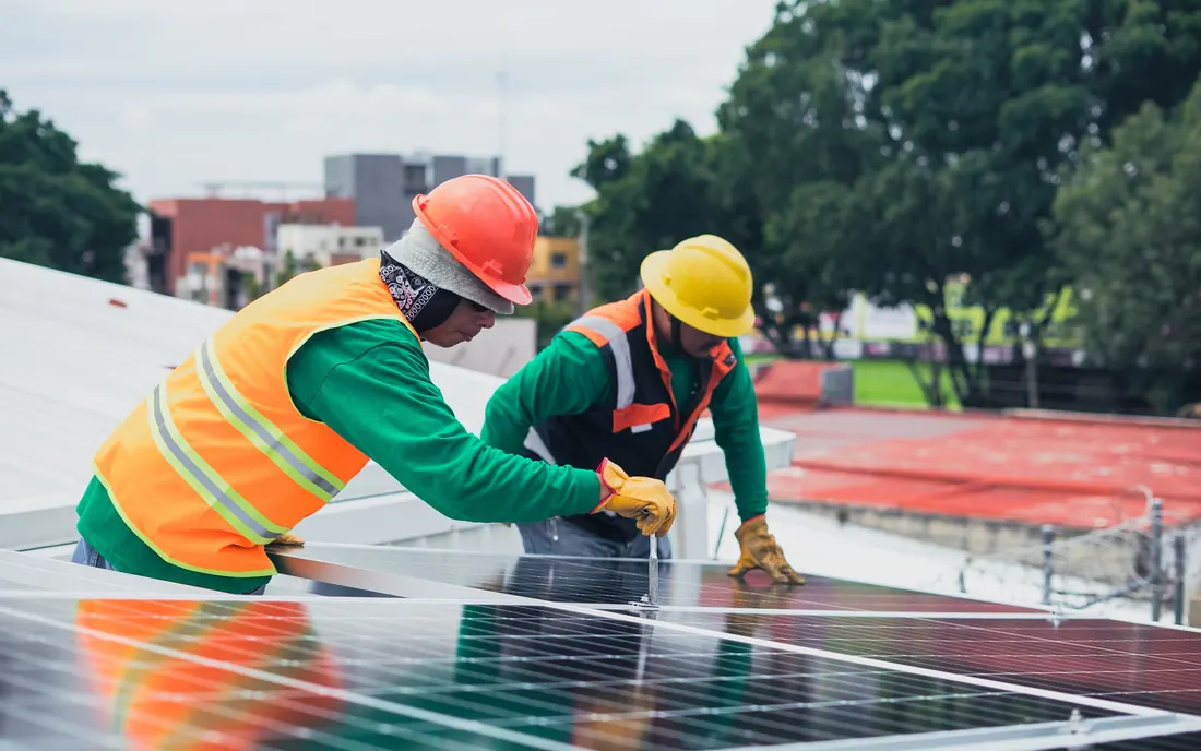 Two solar technicians in hard hats, hi-vis vests and work gloves fastening a solar panel to a metal roof, with trees and low buildings behind them. Photo by Los Muertos Crew on Pexels
