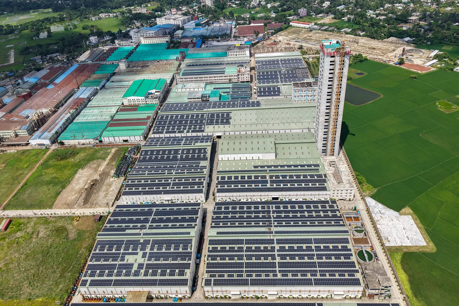 Aerial view of a large industrial complex with long rows of solar panels covering the factory roofs, a tower crane standing beside one building and green fields and a town beyond. Photo by Sarowar Hussain on Pexels
