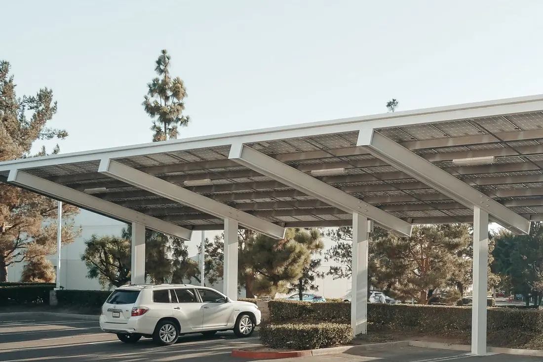 Solar canopy over a commercial parking area with panels on its underside, a white SUV parked beneath it, marked bays and a trimmed hedge behind. Photo by Kindel Media on Pexels