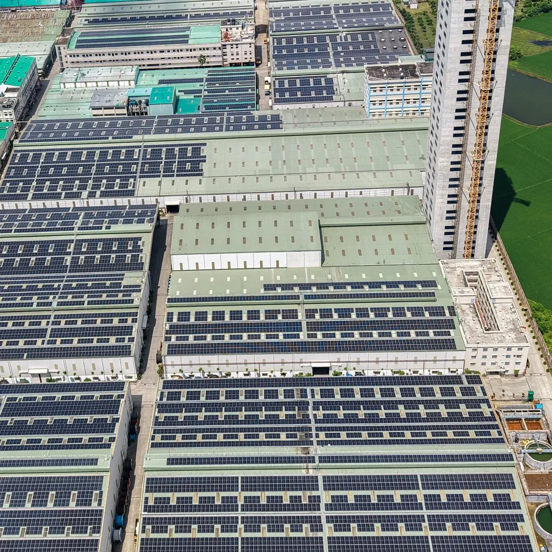 Aerial view of factory roofs covered in rows of solar panels, with a tower crane rising beside one of the buildings. Photo by Sarowar Hussain on Pexels