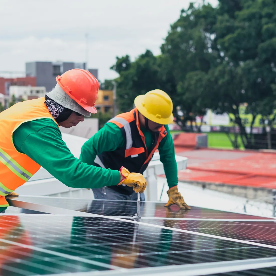 Two technicians in hard hats and hi-vis vests fastening a solar panel on a metal roof, one tightening a bolt with a tool. Photo by Los Muertos Crew on Pexels
