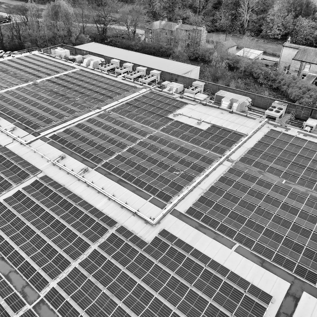 Black and white aerial view of a flat industrial roof covered in rows of solar panels, with rooftop plant units along one edge and trees and houses beyond. Photo by Altaf Shah on Pexels