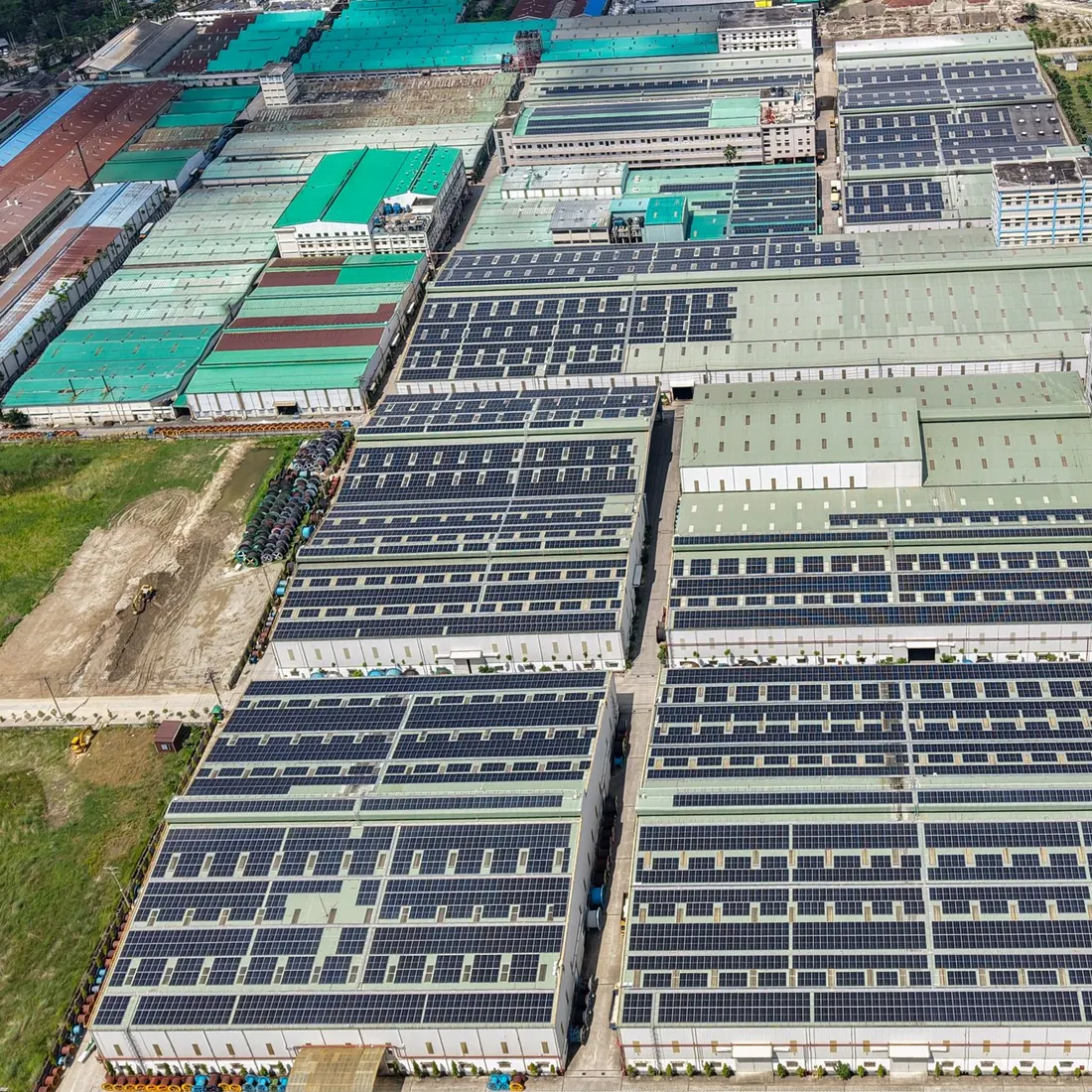 Aerial view of factory roofs covered in rows of solar panels, with a dirt yard, stacked materials and green fields at the edges. Photo by Sarowar Hussain on Pexels