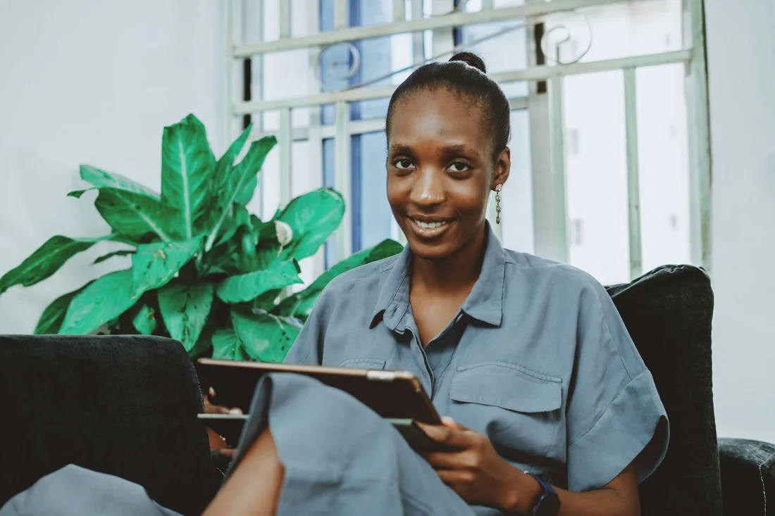 Smiling woman holding a tablet in a bright living room with plants. Photo by David Kwewum on Pexels