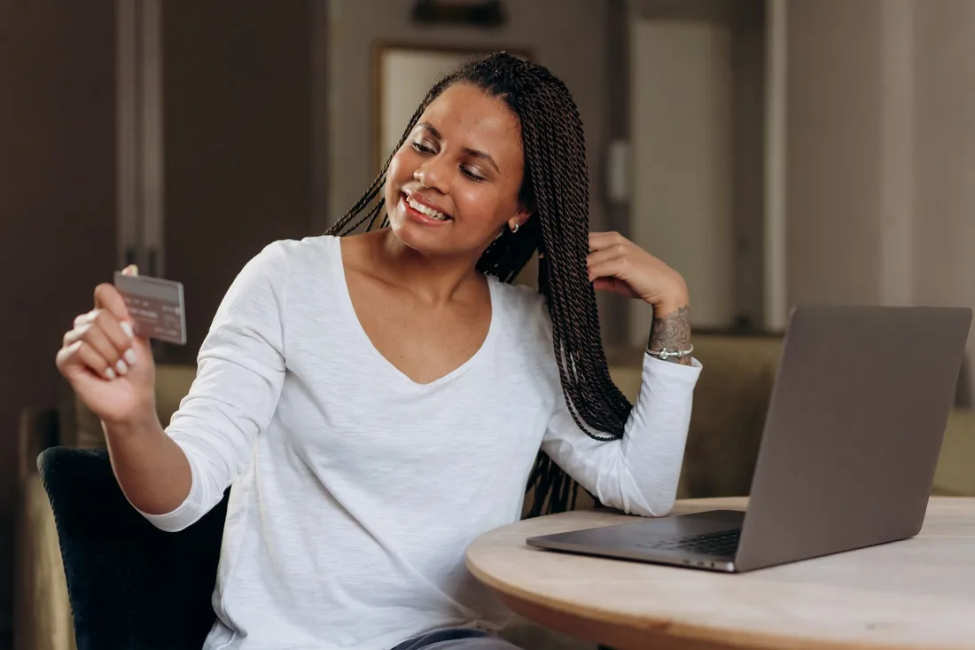 Smiling woman holding a bank card at a table with her laptop. Photo by Mikhail Nilov on Pexels
