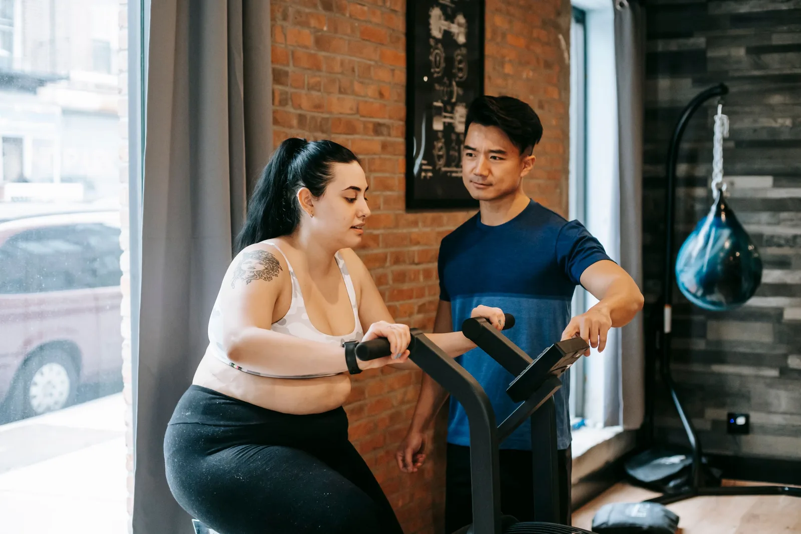 Personal trainer coaching a woman on a gym machine in a bright brick-walled studio. Photo by Andres  Ayrton on Pexels