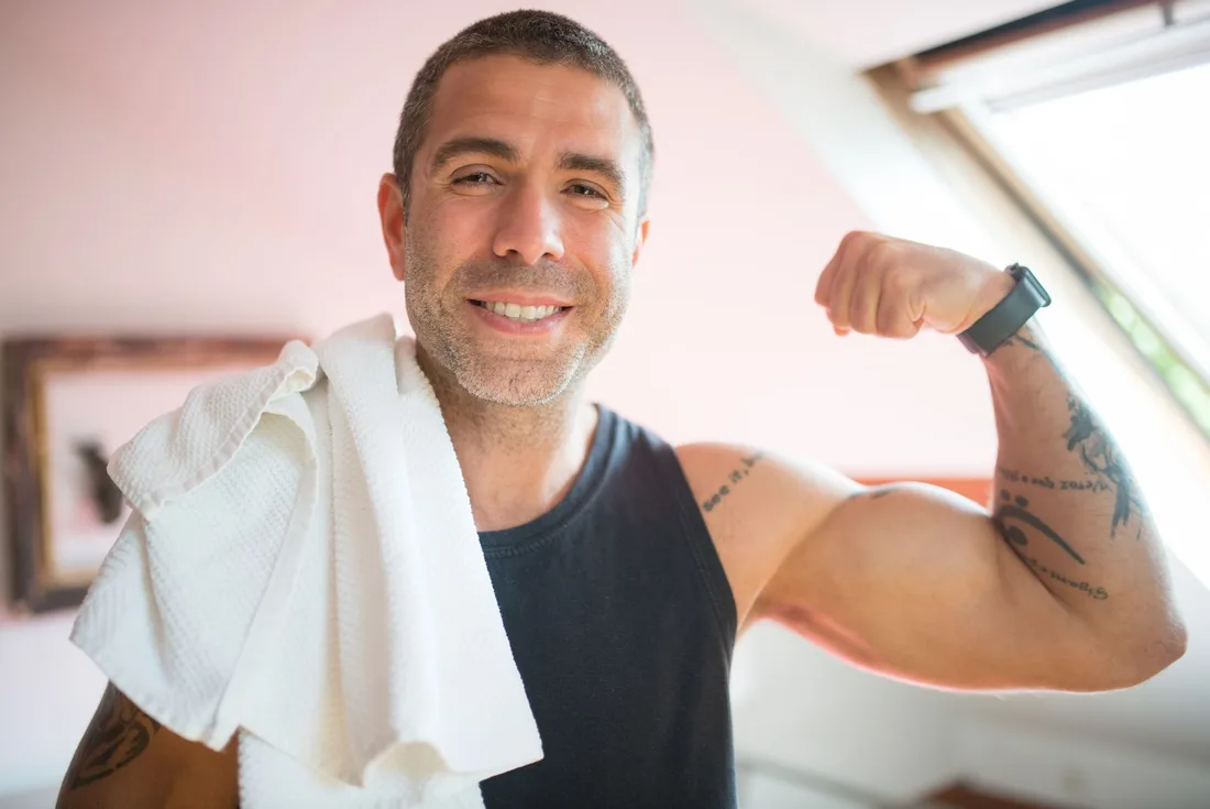 Smiling fit man with a white towel flexing his arm in a bright gym. Photo by Kampus Production on Pexels