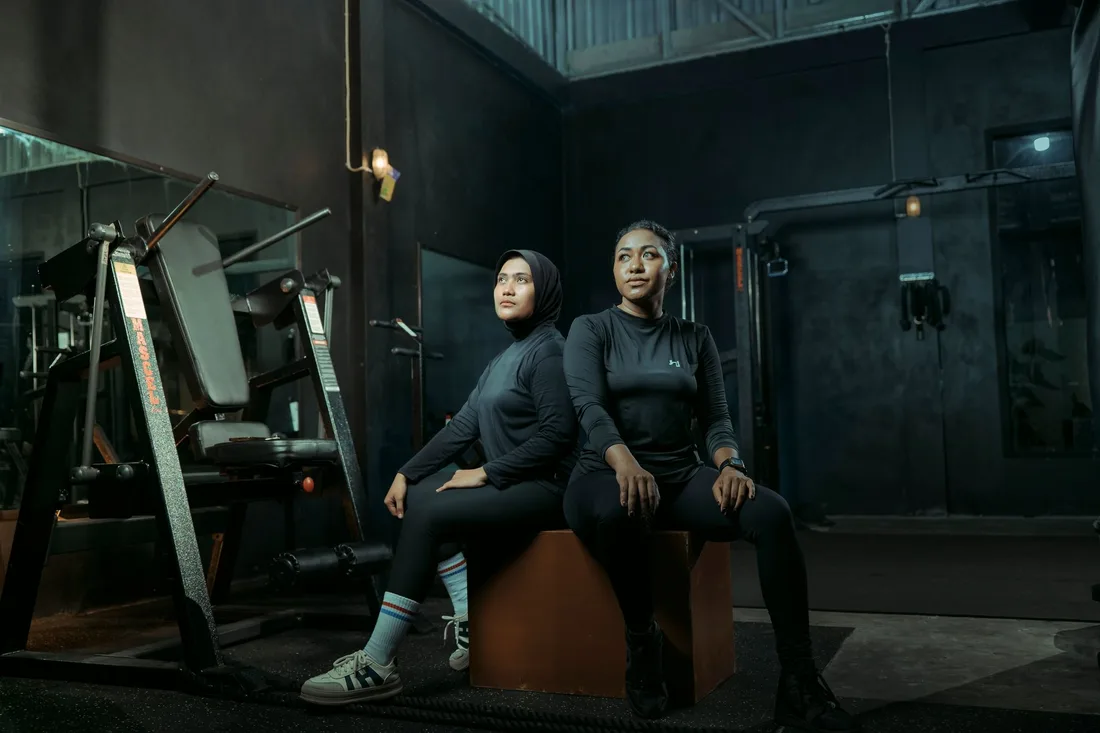 Two women in athletic wear sitting on a box with gym equipment in the background. Photo by Asso Myron on Pexels