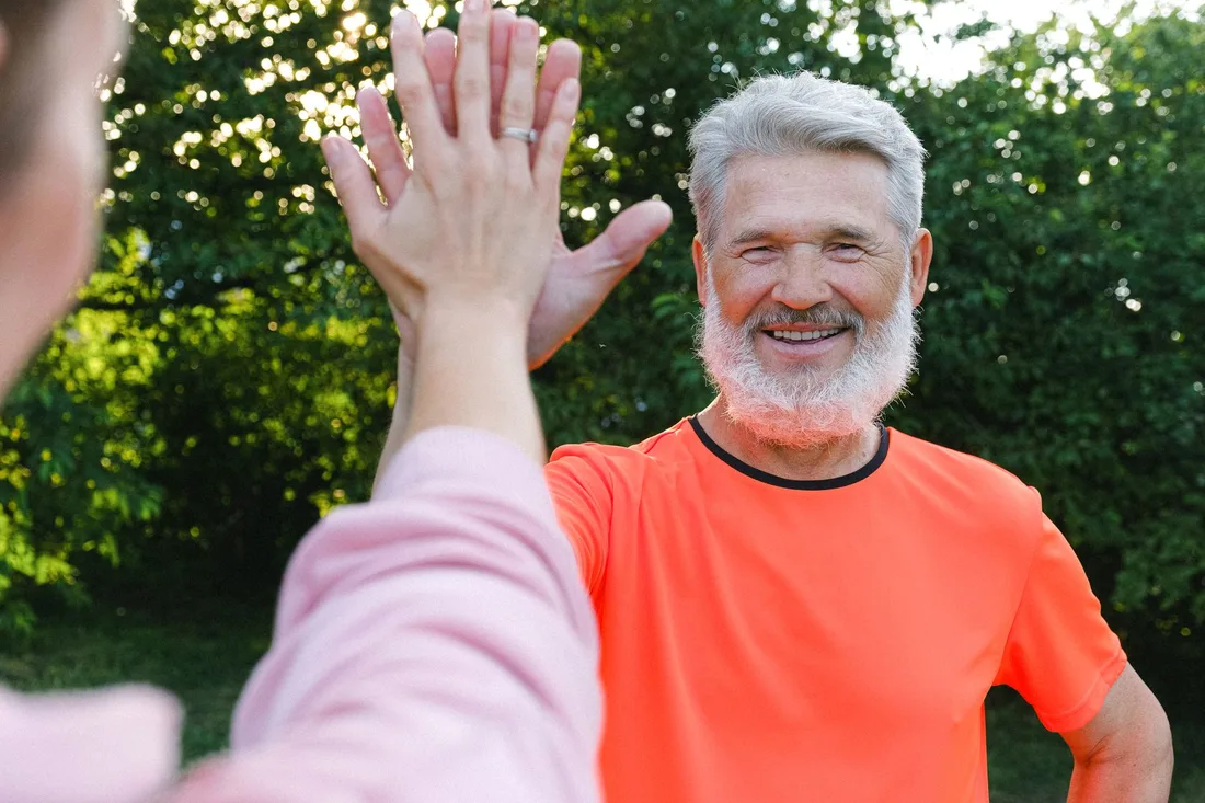 Smiling grey-bearded senior in an orange shirt giving a high five outdoors. Photo by Anna Shvets on Pexels