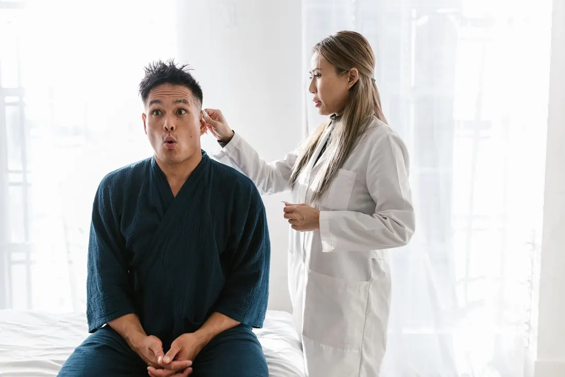 Clinician in a white coat checking the ear of a seated adult man in a dark blue robe, in a bright white room. Photo by RDNE Stock project on Pexels