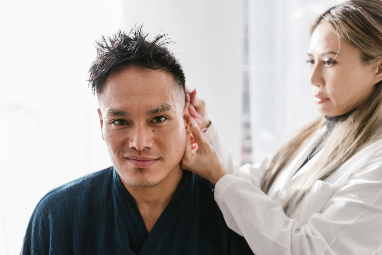 Clinician in a white coat checking the ear of a smiling adult man in a dark blue robe, seated in a bright white room. Photo by RDNE Stock project on Pexels