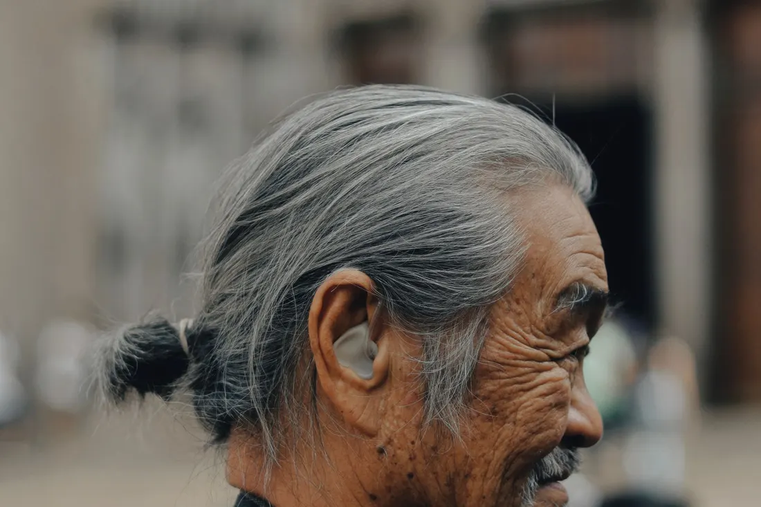 Profile of a senior man with grey hair tied back and a small in-the-ear hearing aid, photographed outdoors against a blurred background. Photo by Thedollasyn on Pexels